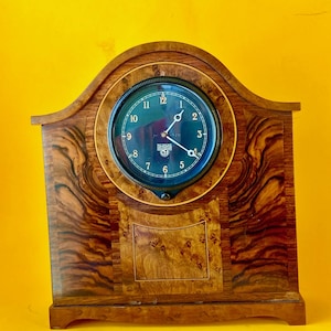 May include: A mantel clock with a wooden case featuring a prominent burl wood grain pattern. The circular clock face is dark with white numerals from 1 to 12 and white hands. The text Smiths is printed on the face below the center point.
