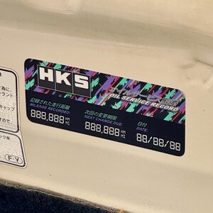 May include: A black and white sticker with the HKS logo and the text "Oil Service Record" in Japanese and English. The sticker also includes the text "Mileage Recorded" and "Next Change Due" with numbers in kilometers and miles. The date is 88/88/88.
