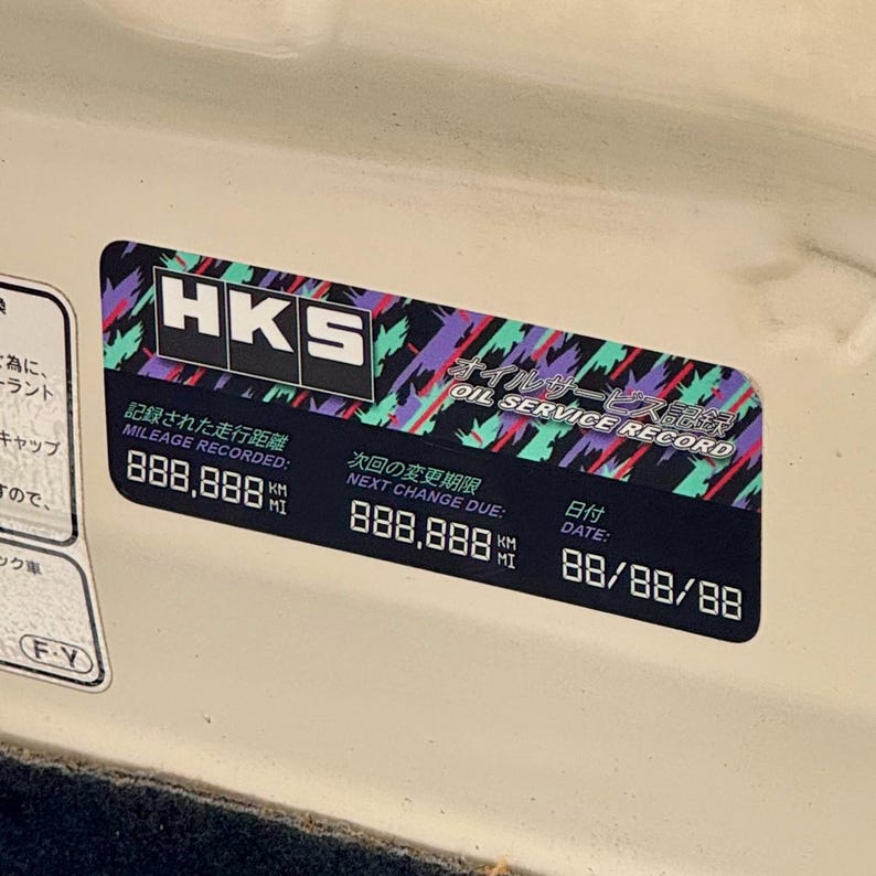 May include: A black and white sticker with the HKS logo and the text "Oil Service Record" in Japanese and English. The sticker also includes the text "Mileage Recorded" and "Next Change Due" with numbers in kilometers and miles. The date is 88/88/88.