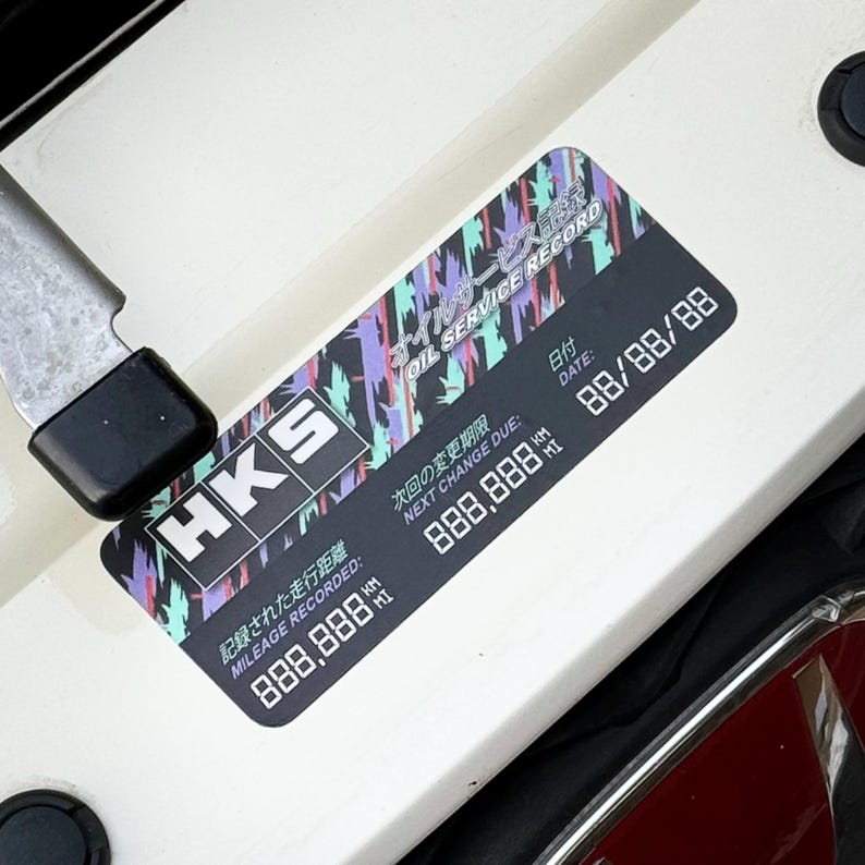 May include: A black and white sticker with the text "HKS" in white letters. The sticker has a colorful abstract design and the text "Oil Service Record" in black letters. The sticker also includes the text "Date", "Mileage Recorded", "Next Change Due", and "888,888 km".