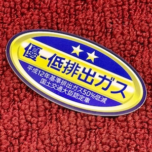 May include: A blue and gold oval sticker with the text "優-低排出ガス" (Yu-Tei Hai Shutsu Gasu) which translates to "Excellent Low Emission Gas". The sticker also includes the text "平成12年基準排出ガス50%低減" (Heisei 12 Nen Kijun Hai Shutsu Gasu 50% Teigen) which translates to "平成12 Year Standard Emission Gas 50% Reduction" and "国土交通大臣認定車" (Kokudo Kotsu Daijin Nintei Sha) which translates to "Ministry of Land, Infrastructure, Transport and Tourism Certified Vehicle".