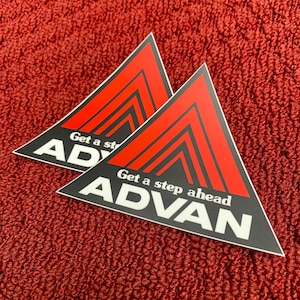 Può includere: Due adesivi neri a forma di triangolo con strisce rosse e testo bianco. Il testo recita "Get a step ahead ADVAN".