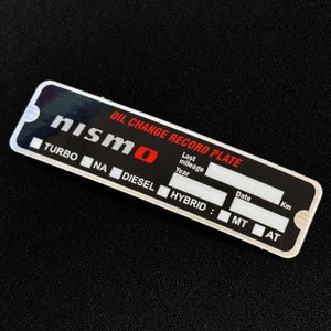 Può includere: Una piastra rettangolare nera e argento per il registro del cambio olio Nismo. La piastra include campi per il chilometraggio, l'anno, la data e il tipo di motore, tra cui Turbo, NA, Diesel e Hybrid. Il testo "OIL CHANGE RECORD PLATE" è stampato in rosso.