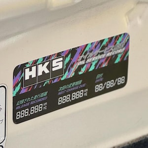 May include: A black and white sticker with the text "HKS Oil Service Record" in a colorful, geometric design. The sticker includes the text "Mileage Recorded: 888,888 km" and "Next Change Due: 888,888 km". The date is "88/88/88".