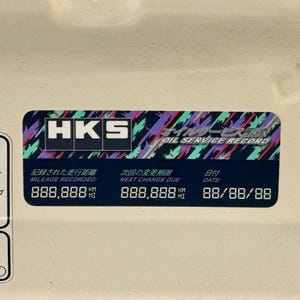 May include: A black and white sticker with the text "HKS Oil Service Record" in a colorful, geometric design. The sticker includes the text "Mileage Recorded: 888,888 km", "Next Change Due: 888,888 km", and "Date: 88/88/88".