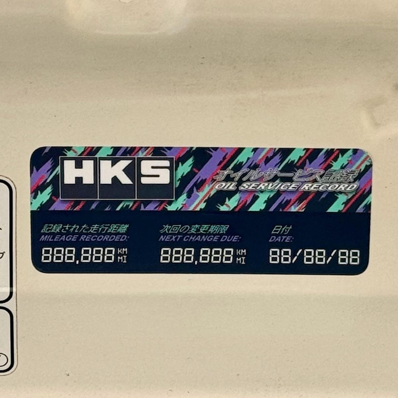 May include: A black and white sticker with the text "HKS Oil Service Record" in a colorful, geometric design. The sticker includes the text "Mileage Recorded: 888,888 km", "Next Change Due: 888,888 km", and "Date: 88/88/88".