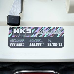 May include: A black and white sticker with the text "HKS Oil Service Record" in a colorful, geometric pattern. The sticker includes the text "Mileage Recorded: 888,888 km", "Next Change Due: 888,888 km", and "Date: 88/88/88".