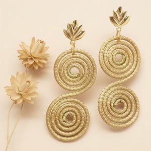 Puede incluir: Pendientes dorados en espiral con adornos en forma de hoja. Los pendientes tienen un diseño circular con una textura enrollada. Se muestran dos pares de pendientes, junto con flores secas sobre un fondo melocotón claro.