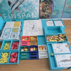 Pode incluir: Caixa de jogo de tabuleiro aberta com o título "FINSPAN: A Wingspan Game". O jogo inclui vários componentes: peças de jogo azuis, amarelas, vermelhas e laranja, cartas e folhas de instruções. A caixa é azul clara.