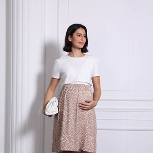 Könnte beinhalten: Eine schwangere Person trägt ein weißes Umstands-Oberteil mit einem braunen und weißen Polka-Dot-Rock. Die Person hält eine weiße Clutch und trägt weiße Sandalen.