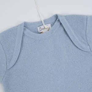 Könnte beinhalten: Ein hellblauer gestrickter Baby-Pullover mit Rundhalsausschnitt und kurzen Ärmeln. Der Pullover hat ein kleines Etikett mit dem Text "Luna" darauf.