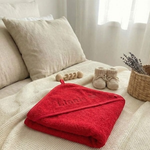Könnte beinhalten: Ein rotes Baby-Badetuch mit Kapuze und dem Namen "Linnie" auf einer cremefarbenen Decke. Daneben befinden sich ein Paar beige gestrickte Babyschuhe, eine Holzrassel und ein kleiner Korb mit Lavendel.