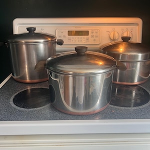 May include: Three stainless steel pots with lids, each featuring dark brown handles and a copper-colored base. The pots are arranged on a stovetop, with one large pot in the front center and two slightly smaller pots positioned behind it.