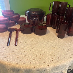 May include: A collection of vintage burgundy-colored kitchenware, including bowls, serving dishes, mugs, and a covered casserole dish. Two serving spoons and a salad fork are also visible. The items are arranged on a white lace tablecloth.