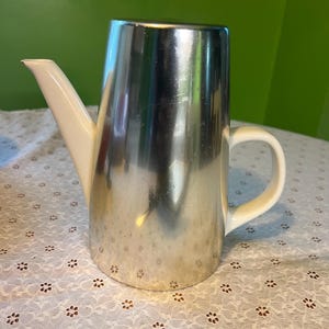 May include: A stainless steel coffee pot with a white spout and handle. The pot has a cylindrical body and a shiny, reflective surface. The spout is angled and the handle is curved.