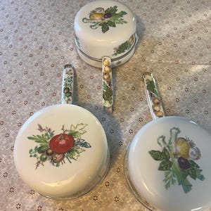 May include: Three white enameled metal saucepans with long handles, each featuring a unique botanical illustration of vegetables. The designs include a red tomato, various onions, and green herbs, with matching vegetable patterns on the handles.