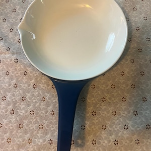 May include: A white enamel pan with a blue handle. The pan has a rounded shape with a pouring spout. The handle is a solid blue color with a hole at the end for hanging. The pan is likely used for cooking.