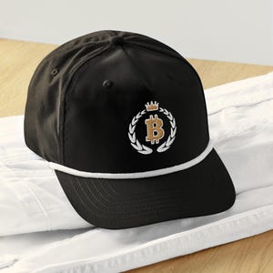 May include: Black baseball cap with a white rope detail and a curved brim. The cap features a Bitcoin symbol in gold, encircled by a laurel wreath and a crown. The design is embroidered on the front.