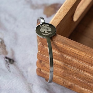 May include: A silver-colored cuff bracelet with the word "FLOURISH" and "PSALM 52:8" engraved on a circular charm. The charm features a tree design. The bracelet is resting on a wooden tray.
