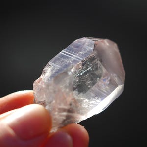 Puede incluir: Un cristal de cuarzo transparente con un tono rosado y una textura natural y rugosa. El cristal est&aacute; facetado y tiene una punta en un extremo.
