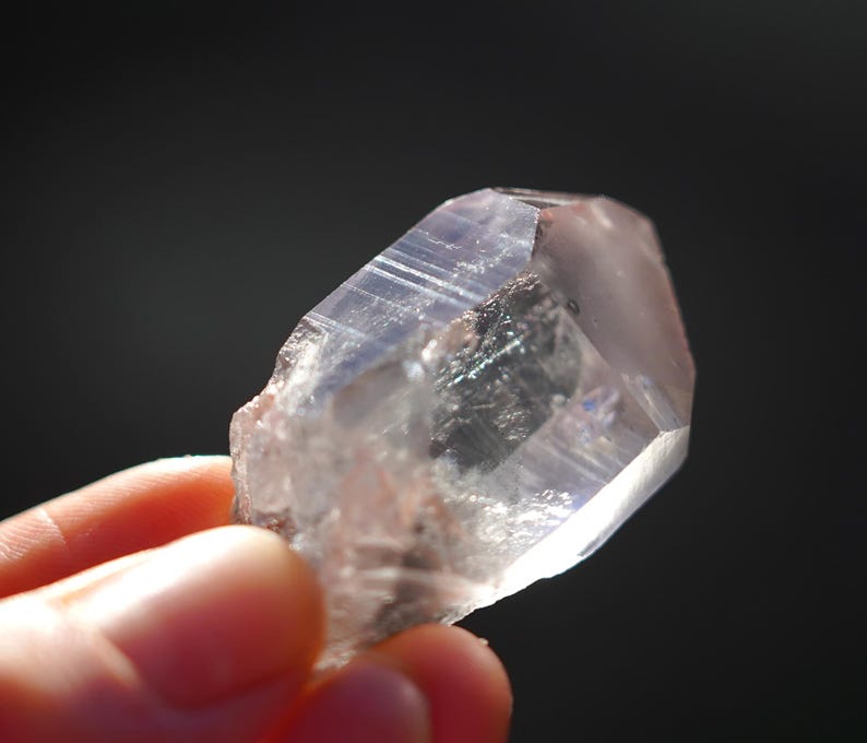 Puede incluir: Un cristal de cuarzo transparente con un tono rosado y una textura natural y rugosa. El cristal est&aacute; facetado y tiene una punta en un extremo.