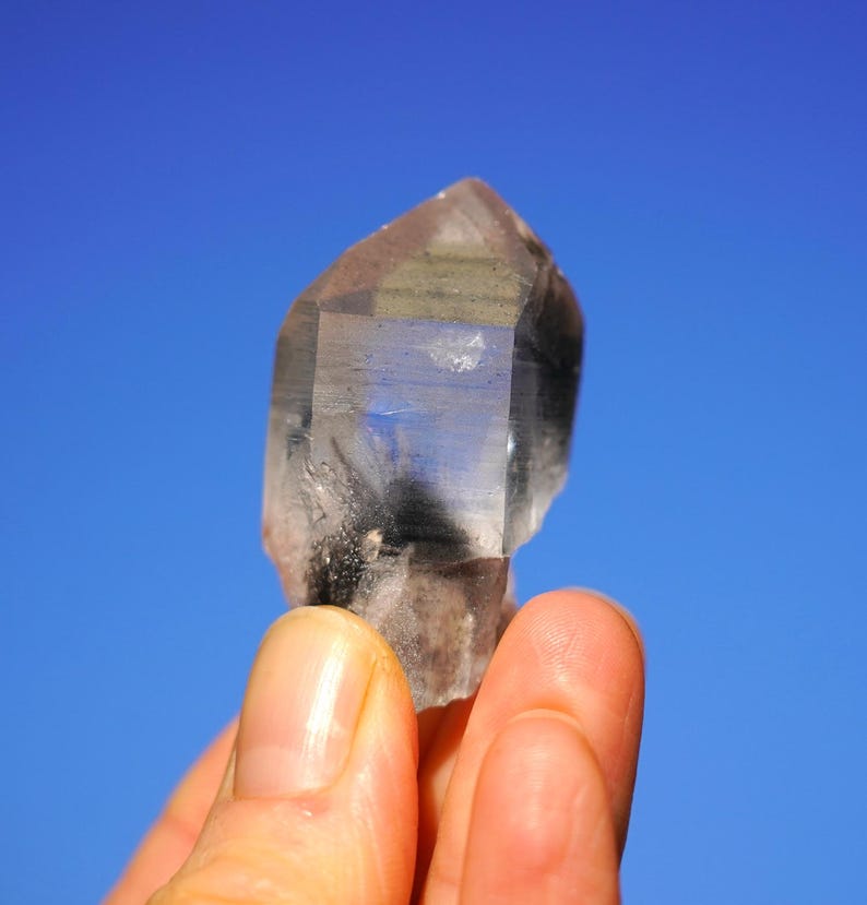 Puede incluir: Un cristal de cuarzo transparente con una inclusi&oacute;n gris ahumada, sostenido en una mano contra un cielo azul brillante.