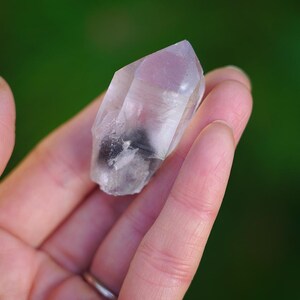 Puede incluir: Un cristal de cuarzo transparente con un toque de rosa y una inclusi&oacute;n negra. El cristal es puntiagudo en un extremo y tiene una textura natural y rugosa.