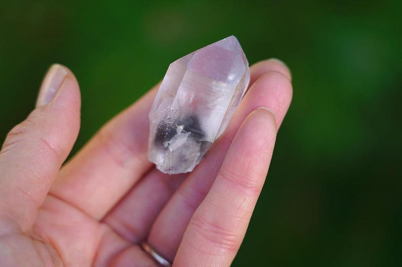 Puede incluir: Un cristal de cuarzo transparente con un toque de rosa y una inclusi&oacute;n negra. El cristal es puntiagudo en un extremo y tiene una textura natural y rugosa.