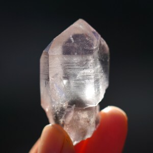 Puede incluir: Un cristal transparente y facetado con un tono rosa claro. El cristal tiene forma de punta y una superficie lisa y pulida.