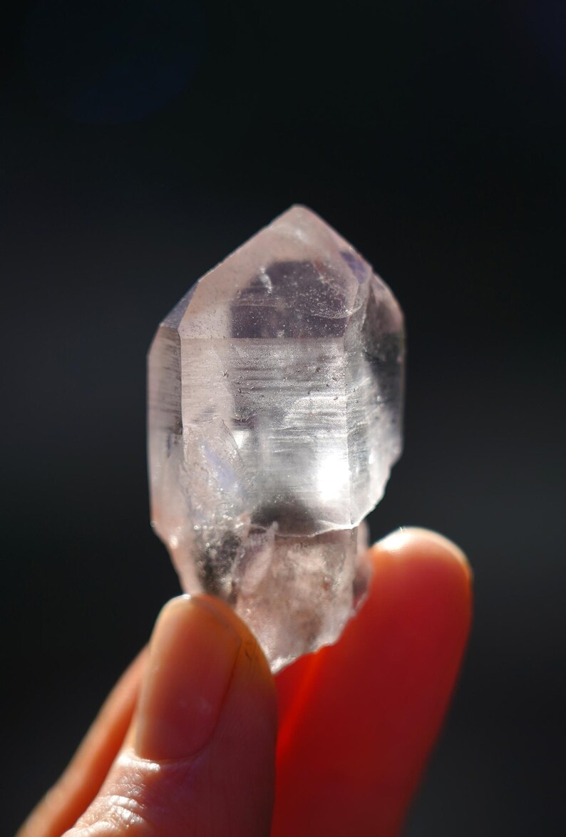 Puede incluir: Un cristal transparente y facetado con un tono rosa claro. El cristal tiene forma de punta y una superficie lisa y pulida.