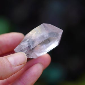 Puede incluir: Un cristal transparente y facetado con un ligero brillo iridiscente. El cristal se sostiene en una mano, mostrando su forma y claridad.
