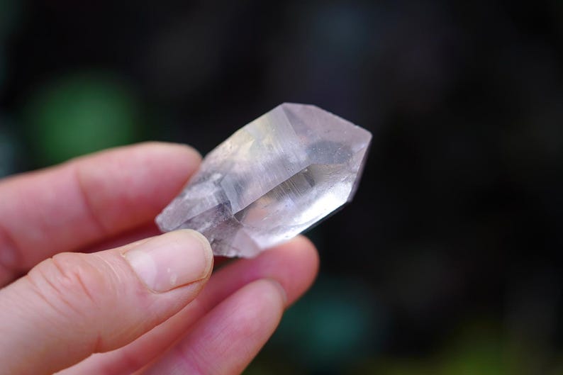 Puede incluir: Un cristal transparente y facetado con un ligero brillo iridiscente. El cristal se sostiene en una mano, mostrando su forma y claridad.