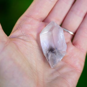 Puede incluir: Un cristal de cuarzo transparente con un ligero tono rosado y una inclusi&oacute;n gris nublada. El cristal tiene forma de punta de doble extremo y se sostiene en una mano.