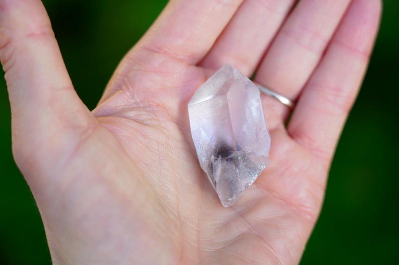 Puede incluir: Un cristal de cuarzo transparente con un ligero tono rosado y una inclusi&oacute;n gris nublada. El cristal tiene forma de punta de doble extremo y se sostiene en una mano.