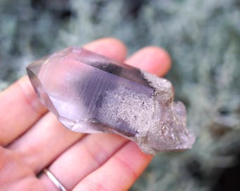 Puede incluir: Un cristal de cuarzo transparente con una inclusión gris ahumada. El cristal es puntiagudo en un extremo y tiene una superficie rugosa y texturizada.