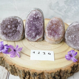 Könnte beinhalten: Vier Amethyst-Kristall-Geoden, jede in einer anderen Größe und Form, werden auf einer Holzscheibe präsentiert. Die Geoden sind hellviolett und haben eine raue, natürliche Textur. Die Holzscheibe ist hellbraun und von lila Blüten umgeben.