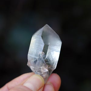 Puede incluir: Un cristal de cuarzo transparente con doble terminaci&oacute;n, tambi&eacute;n conocido como cristal de doble punta. El cristal se sostiene en una mano, mostrando su claridad y facetas.