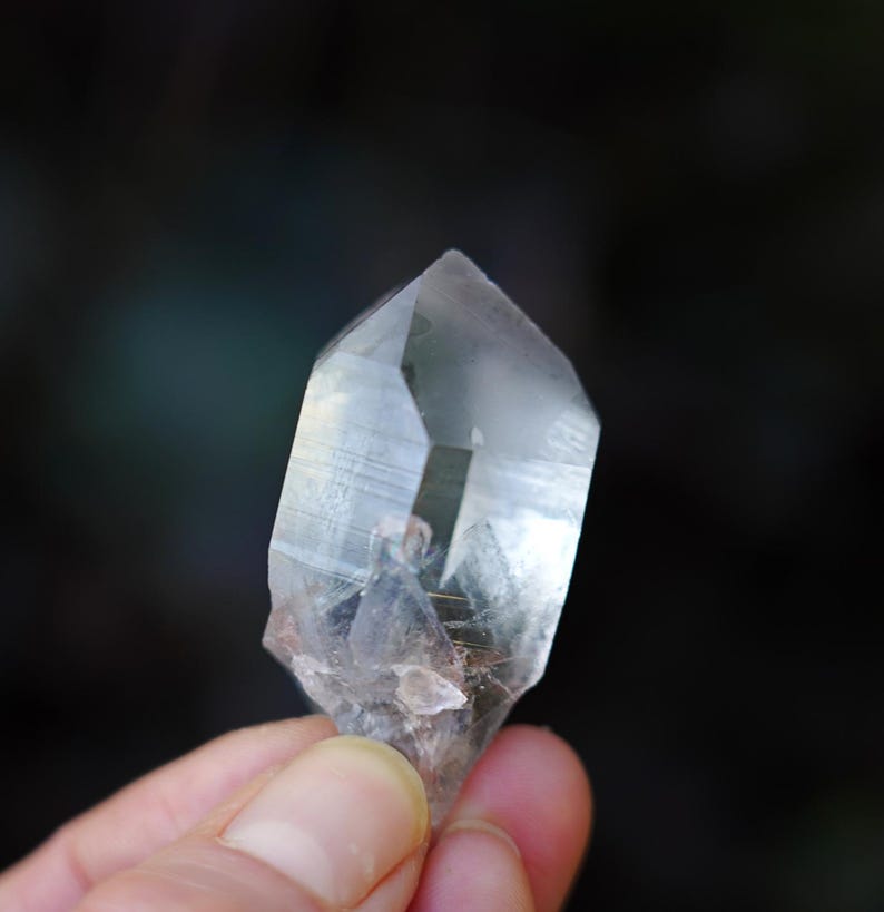 Puede incluir: Un cristal de cuarzo transparente con doble terminaci&oacute;n, tambi&eacute;n conocido como cristal de doble punta. El cristal se sostiene en una mano, mostrando su claridad y facetas.