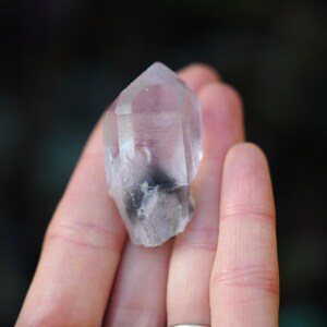 Puede incluir: Una punta de cristal de cuarzo transparente con una inclusi&oacute;n gris. El cristal se sostiene en una mano.