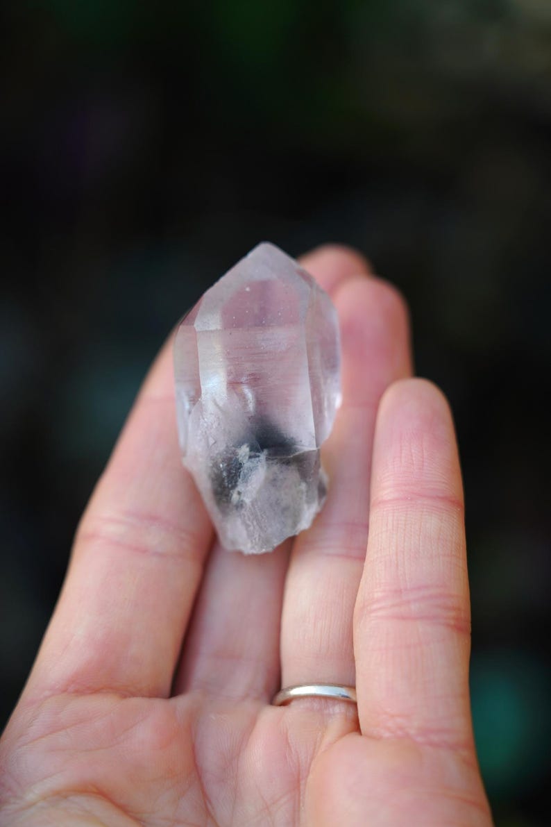 Puede incluir: Una punta de cristal de cuarzo transparente con una inclusi&oacute;n gris. El cristal se sostiene en una mano.