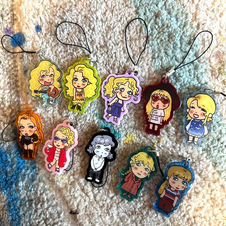 May include: A collection of colorful acrylic charms featuring cartoon illustrations of women in various outfits. Each charm has a black loop for attachment. The charms are in various colors, including yellow, orange, pink, purple, red, and blue.