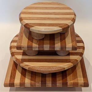 May include: A set of three wooden risers in different shapes and sizes. The risers are made of light and dark brown wood, with a striped pattern on the surfaces. Each riser has rounded edges and small, round feet.