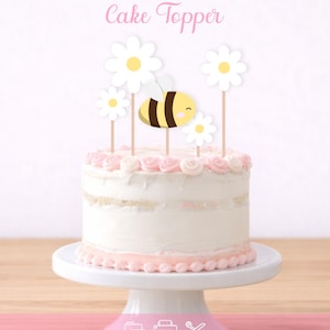 Puede incluir: Una tarta blanca con glaseado rosa y un adorno para tarta de abeja y margarita. La tarta se encuentra sobre un soporte blanco para tartas. Las palabras "Cake Topper" están escritas en rosa en la parte superior de la imagen.