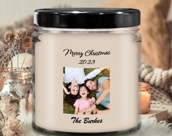 May include: A clear glass candle jar with a black lid. The candle features a family photo and the text "Merry Christmas 2023" and "The Burkes". The candle is surrounded by a cozy, neutral-toned setting.
