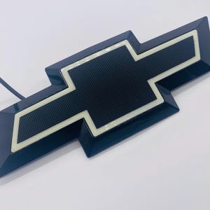 Puede incluir: Un emblema de Chevrolet en forma de pajarita de color azul oscuro con un centro negro texturizado rodeado por un borde blanco brillante. El emblema tiene una forma geométrica e incluye un cable de alimentación azul fino en el lado izquierdo.