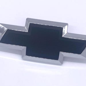 May include: A Chevrolet bowtie emblem featuring a polished chrome border surrounding a dark blue, textured centre section. The iconic cross-shaped logo has sharp, angular edges and a reflective metallic finish on the outer frame.