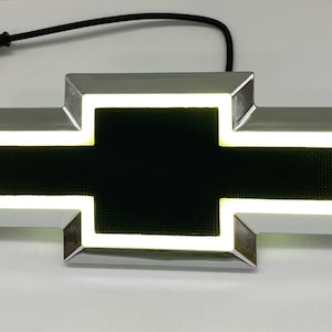 Puede incluir: Un emblema de Chevrolet en forma de pajarita con un borde de cromo pulido y un centro negro texturizado. El emblema está iluminado por una tira de luz LED blanca brillante que perfila la forma interior del logotipo. Un cable eléctrico negro sobresale.