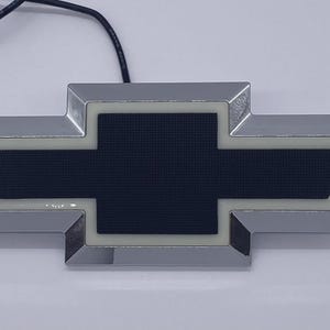 Puede incluir: Un emblema de Chevrolet en forma de pajarita con un borde cromado pulido, un contorno iluminado en blanco y un panel central oscuro texturizado. El emblema incluye un cable eléctrico negro fino que sale del borde superior para conectar.
