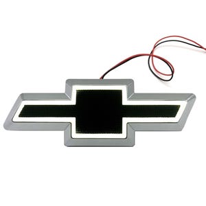 May include: A chrome-finished Chevrolet bowtie emblem featuring a black textured centre panel outlined by a bright white LED light strip. The automotive badge includes two thin red and black electrical wires extending from the top edge.