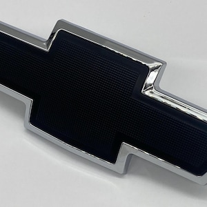 May include: A Chevrolet bowtie emblem featuring a polished chrome border surrounding a textured matte black centre. The iconic cross-shaped logo has a distinct geometric design with a raised metallic edge and a recessed, grid-patterned black inlay.